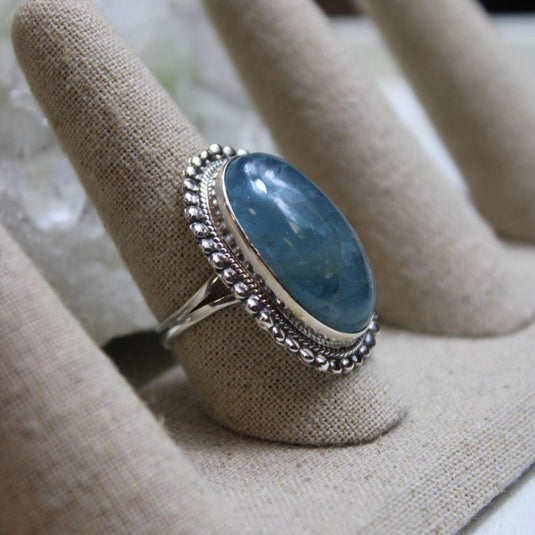 A large oval Aquamarine gemstone set in a silver ring with a beaded border. The ring is displayed on a ring holder with other rings.