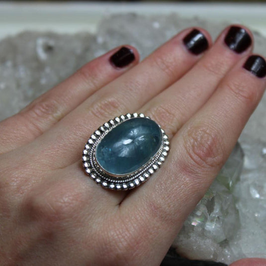 A woman's hand wearing a large oval Aquamarine ring set in silver with a beaded border.