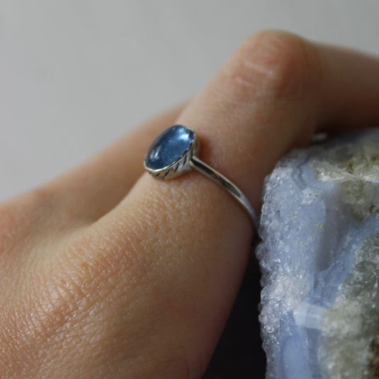 An oval-shaped aquamarine gemstone set in a sterling silver ring, shown on a hand against a blue background.