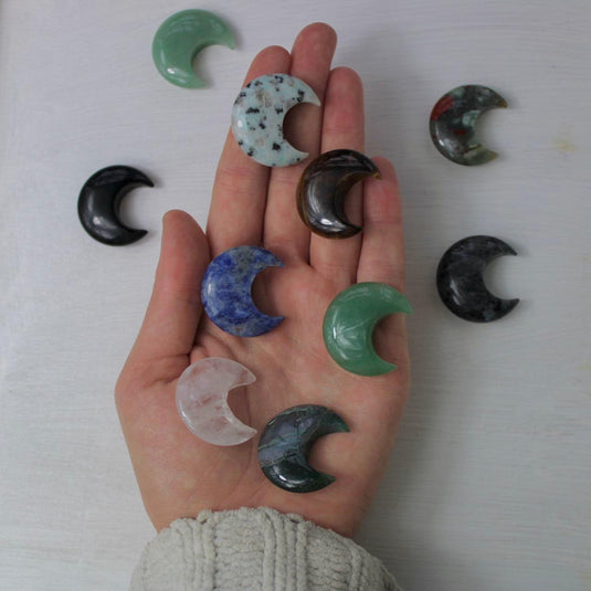 A hand holding a variety of crescent moon-shaped crystal gemstones, including green, blue, brown, black, and clear quartz.