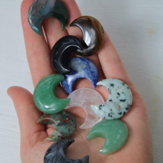 A hand holding a variety of crescent moon-shaped crystal gemstones, including green, blue, brown, black, and clear quartz.