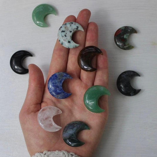 A hand holding a variety of crescent moon-shaped crystal gemstones, including green, blue, brown, black, and clear quartz.