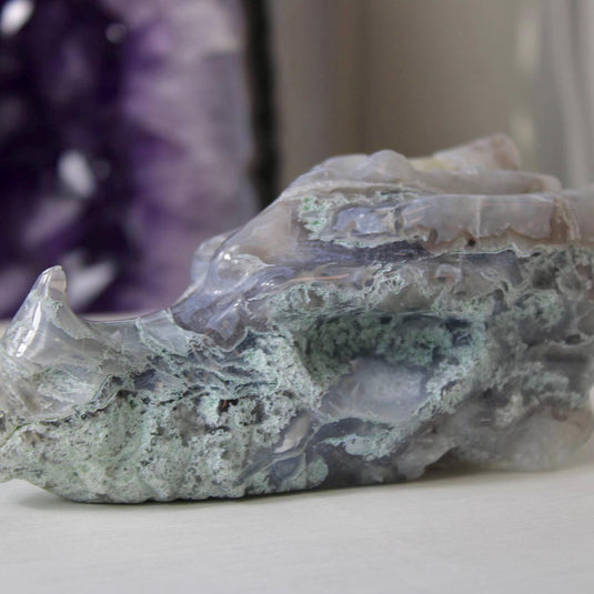 A hand-carved moss agate dragon head, showing intricate details like horns, teeth, and scales. The stone has a beautiful blend of blue, purple, and green hues.