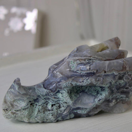 A hand-carved moss agate dragon head, showing intricate details like horns, teeth, and scales. The stone has a beautiful blend of blue, purple, and green hues.