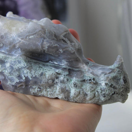 A hand-carved moss agate dragon head, showing intricate details like horns, teeth, and scales. The stone has a beautiful blend of blue, purple, and green hues.