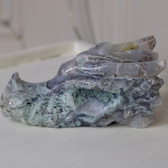 A hand-carved moss agate dragon head, showing intricate details like horns, teeth, and scales. The stone has a beautiful blend of blue, purple, and green hues.