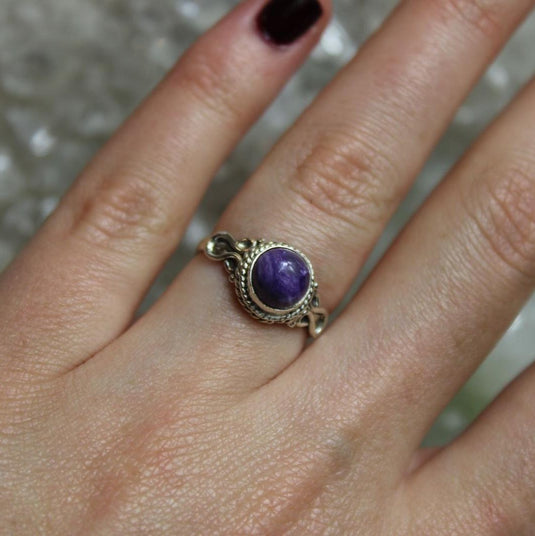 A woman's hand wearing a sterling silver ring with a round, purple Charoite gemstone. The ring has a twisted band and is set against a crystal background.