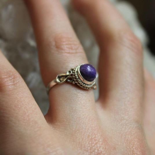 Close-up of a hand wearing a silver ring with a purple Charoite stone. The ring is set on a clear quartz crystal.