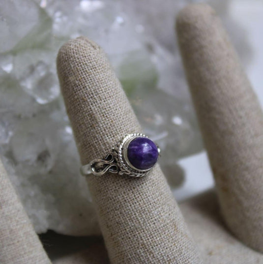 A sterling silver ring with a round, purple Charoite gemstone displayed on a fabric ring cone. The ring cone is placed on a bed of white crystals.