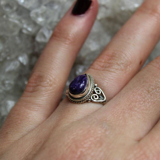 A beautiful charoite ring with a tear drop-shaped cut stone set in a silver band with intricate details.