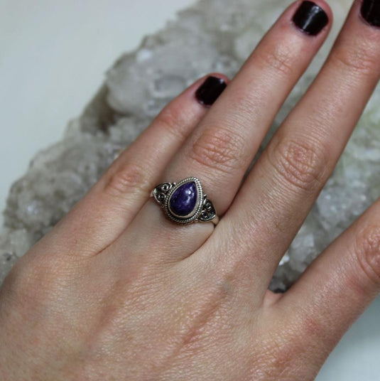 A hand wearing a silver ring with a tear drop-shaped charoite gemstone. The ring is resting on a bed of white crystals.