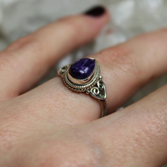 A close-up photo of a tear drop-shaped charoite ring with a silver band. The ring is worn on a finger with dark nail polish.