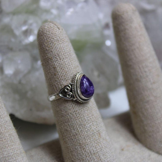 A close-up photo of a tear drop-shaped charoite ring with a silver band on a fabric display.