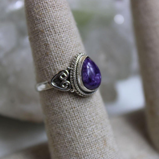 A silver ring with a tear drop-shaped charoite gemstone on a display finger.