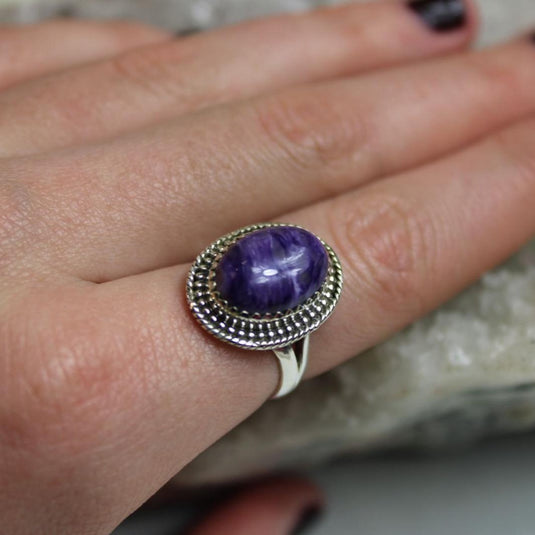 A close-up of a woman's hand wearing a beautiful Charoite ring with a large oval cut stone.