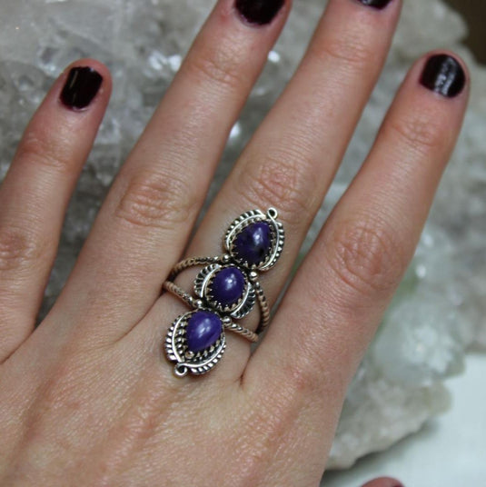 A hand wearing a silver ring with three oval-shaped purple charoite gemstones set in intricate silver filigree.