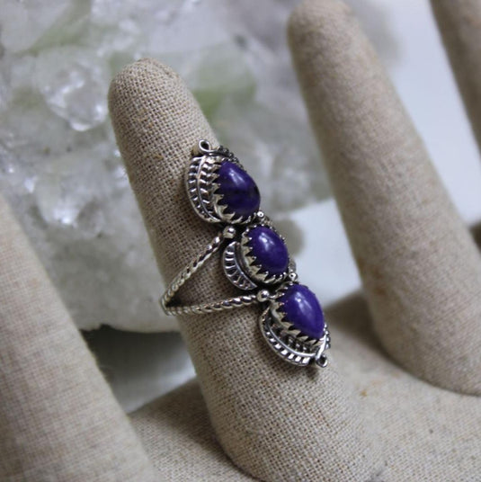 A silver ring with three oval-shaped purple charoite gemstones set in intricate silver filigree, displayed on a ring mandrel.