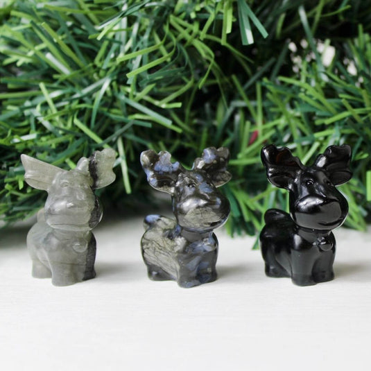 A trio of reindeer figurines carved from various crystals. The leftmost reindeer is made of labradorite, displaying a shimmering iridescent sheen. The middle reindeer is carved from que sera, known for its calming energy and unique patterns. The rightmost reindeer is made of black obsidian, a grounding stone with a deep, reflective surface.