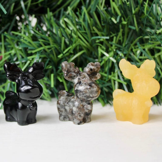 A trio of reindeer figurines, each carved from a different crystal. The leftmost reindeer is made of black obsidian, known for its grounding properties. The middle reindeer is carved from yooperlite, symbolizing strength and stability. The rightmost reindeer is made of orange calcite, a crystal associated with balance and joy.