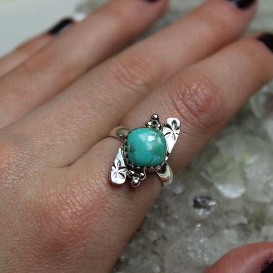 A close up of a sterling silver ring with turquoise gemstone and flower designs displayed on a hand  