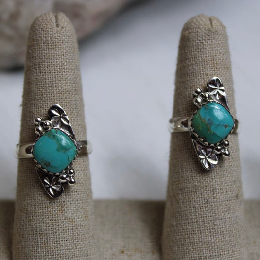 A close up of two sterling silver rings with turquoise gemstones and flower designs, displayed on ring stands