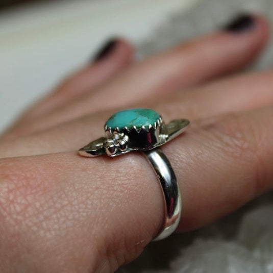 A close up of a sterling silver ring with turquoise gemstone and flower designs displayed on a hand from a side angle 