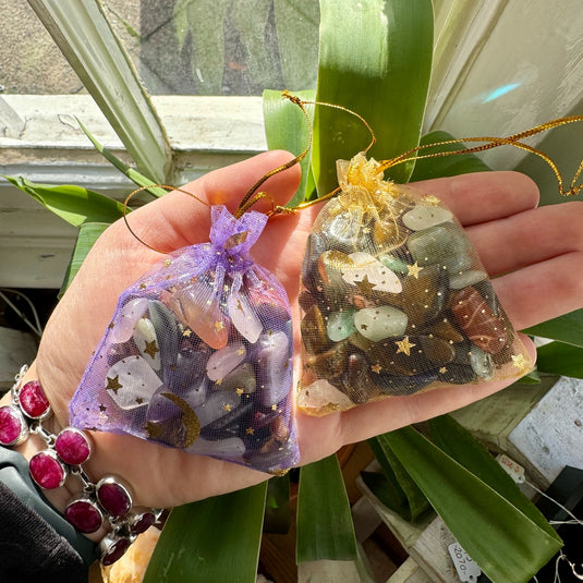 A person's hand holding a handful of small, colourful stones. The stones are various shapes and sizes, and include colours such as purple, green, red, black, and white. The background shows a basket of flowers, a crystal cluster, and other stones.