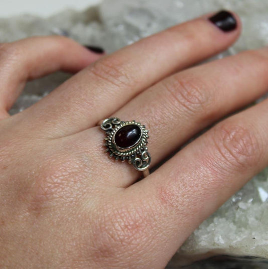 A close-up of a hand wearing a sterling silver ring with a garnet stone. The ring has intricate filigree detailing around the stone.