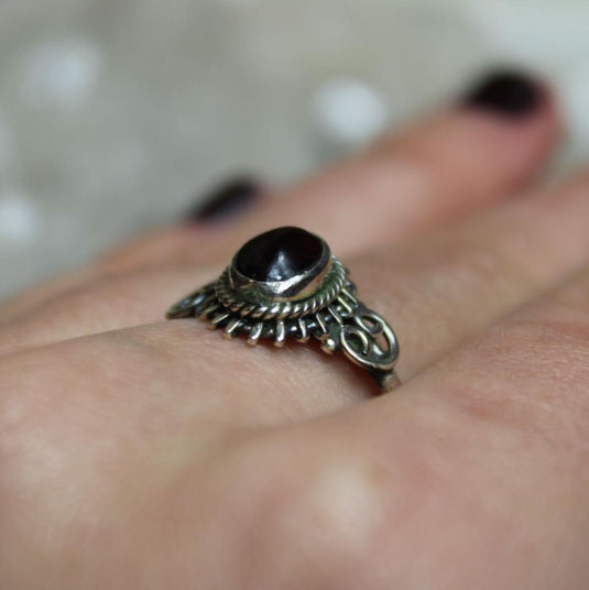 A close-up of a hand wearing a sterling silver ring with a garnet stone. The ring has intricate filigree detailing around the stone.