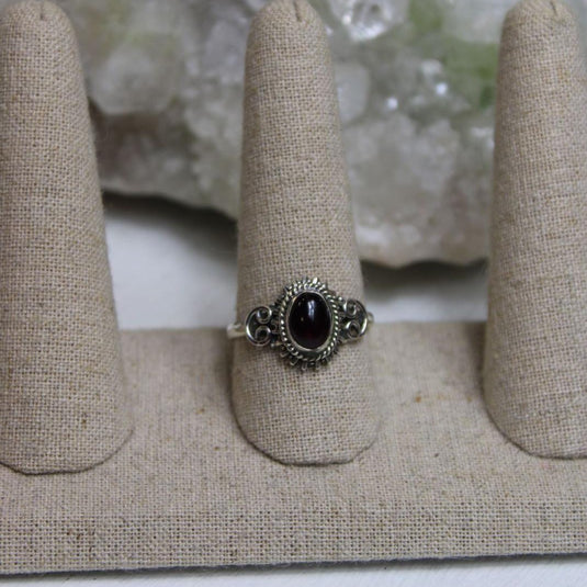 A sterling silver ring with a red garnet stone set in a filigree design. The ring is displayed on a fabric-covered ring holder.