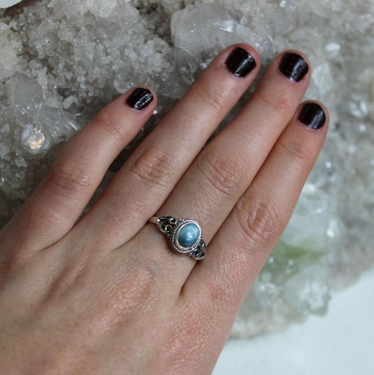 A hand with black nail polish wearing a silver ring with a blue Larimar gemstone, resting on a large crystal.